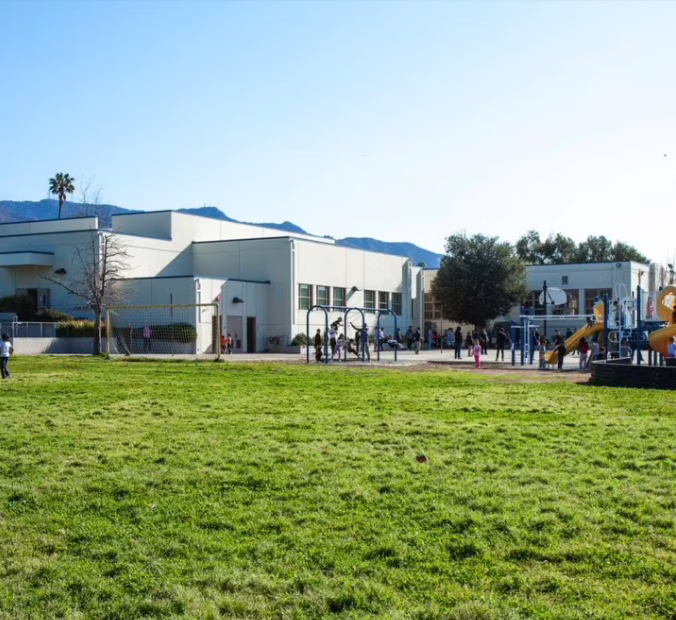 A grassy field leads to a school building with students playing safely on the playground and walking near the entrance, reflecting Safe Schools initiatives. Trees and mountains are visible in the background under a clear blue sky.