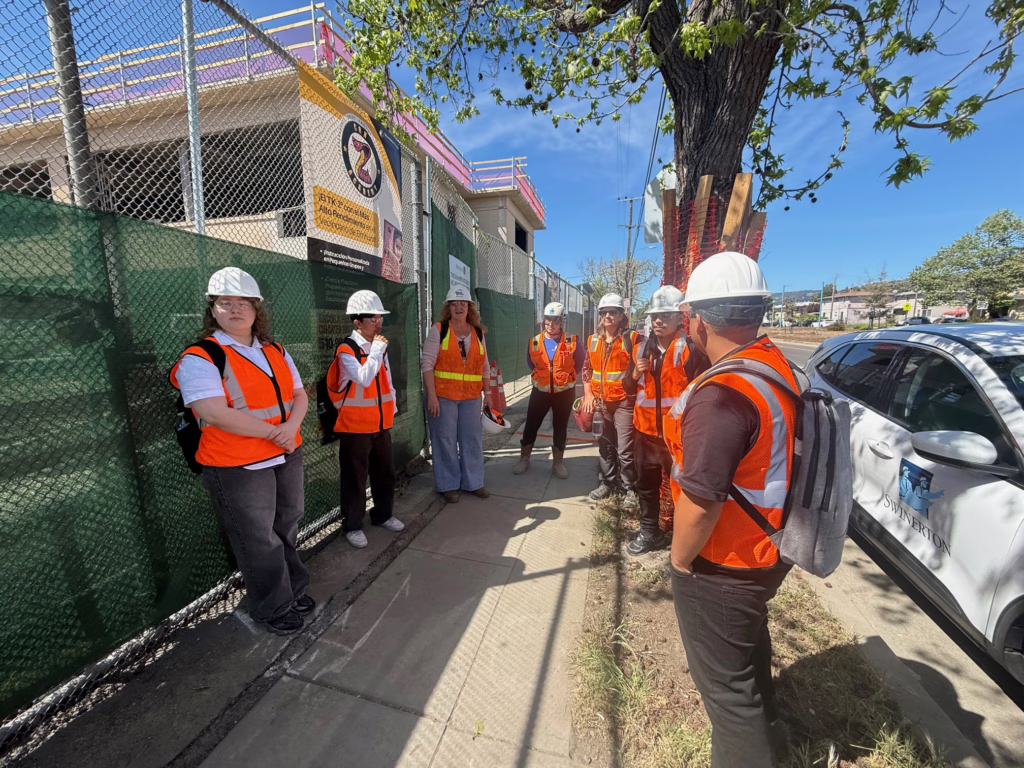 A group of people wearing orange safety vests and white hard hats stand on a sidewalk near a Swinerton construction site, listening to a woman speaking. A fence, signs, and trees are visible in the background—a day in the life on site.