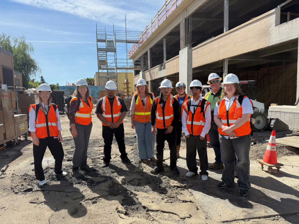 A group of people wearing orange safety vests and white hard hats stand together at a Swinerton construction site, with scaffolding and a partially built structure in the background under a sunny sky.